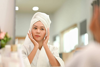 很高兴年轻的女人白色浴袍应用软化保湿脸奶油皮肤脸护理前面镜子