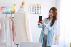 年轻的亚洲女时尚设计师采取照片西装产品智能手机设计工作室