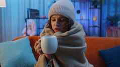 生病的不健康的女人穿他包装格子坐着瑟瑟发抖冷沙发喝热茶