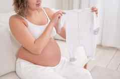 怀孕了女人坐在白色沙发持有衣服预期婴儿