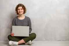 快乐年轻的女人坐着地板上交叉腿移动PC灰色的背景