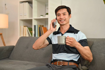 微笑亚洲男人。自由职业者电话谈话喝沙发上舒适的生活房间