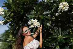 年轻的美丽的女人浪漫的衣服花打印太阳镜珍珠项链手镯背景热带叶子