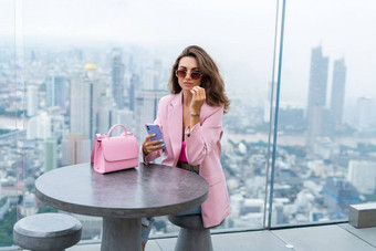 时尚的女人美丽的时尚粉红色的夹克可爱的袋持有移动电话屋顶阳台令人惊异的曼谷视图高地板上工业城市