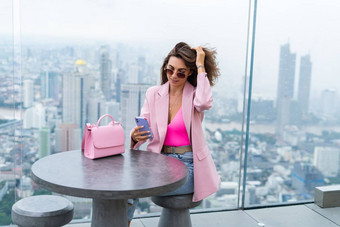 时尚的女人美丽的时尚粉红色的夹克可爱的袋持有移动电话屋顶阳台令人惊异的曼谷视图高地板上工业城市