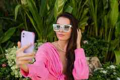 年轻的美丽的浅黑肤色的女人粉红色的衬衫脖子珠宝项链时尚的太阳镜坐在背景热带叶子需要自拍电话