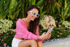 年轻的美丽的浅黑肤色的女人女人粉红色的衬衫珠宝脖子时尚的太阳镜坐在背景热带叶子电话屏幕微笑