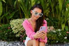 年轻的美丽的浅黑肤色的女人女人粉红色的衬衫珠宝脖子时尚的太阳镜坐在背景热带叶子电话屏幕微笑