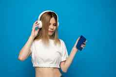 美丽的有吸引力的年轻的金发碧眼的女人穿白色t恤眼镜白色耳机听音乐智能手机跳舞笑蓝色的背景工作室生活方式