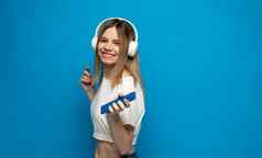 美丽的有吸引力的年轻的金发碧眼的女人穿白色t恤眼镜白色耳机听音乐智能手机跳舞笑蓝色的背景工作室生活方式