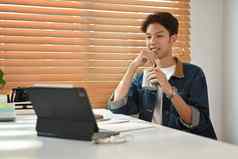 很高兴年轻的亚洲男人。牛仔裤夹克喝咖啡阅读在线新闻数字平板电脑首页办公室