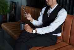 肖像不错的谨慎的亚洲男人。坐着沙发智能手机喝咖啡