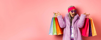 时尚的亚洲高级女人购物穿时尚的衣服太阳镜持有商店袋礼物治疗概念粉红色的背景