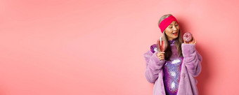 花俏的高级女人有趣的吃甜甜圈喝粉红色的香槟站紫色的人造皮毛外套闪闪发光的衣服工作室背景