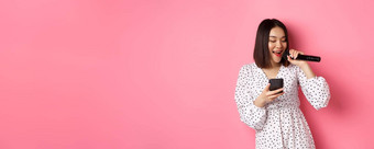 可爱的亚洲女人阅读<strong>歌词</strong>智能手机唱歌麦克风站时尚的衣服粉红色的背景