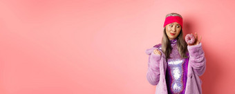 有趣的亚洲高级女人闪闪发光的迪斯科衣服人造皮毛外套诱惑美味的甜甜圈想要吃甜蜜的站粉红色的背景