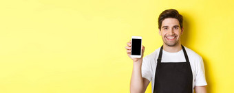 特写镜头英俊的服务员黑色的围裙显示智能手机屏幕推荐应用程序站黄色的背景