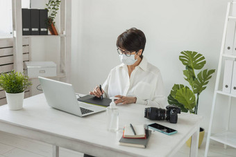 中间岁的女人医疗脸面具图形设计师摄影师数字图形平板电脑工作现代办公室专业女修图编辑坐着现代工作空间通用的设计平板电脑照片相机科维德流感大流行