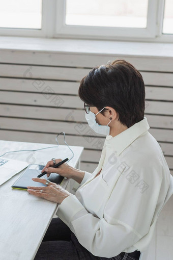 中间岁的女人医疗脸面具图形设计师摄影师数字图形平板电脑工作现代办公室专业女修图编辑坐着现代工作空间通用的设计平板电脑照片相机科维德流感大流行