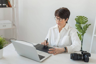 中间岁的女人图形设计师摄影师数字图形平板电脑工作现代办公室专业女修图编辑坐着现代工作空间通用的设计平板电脑照片相机