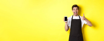 快乐咖啡师黑色的围裙显示智能手机屏幕使翘拇指推荐咖啡馆应用程序站黄色的背景