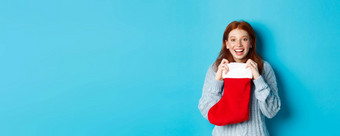 冬天假期礼物概念快乐十几岁的红色头发的人女孩接收圣诞节礼物开放圣诞节长袜微笑惊讶站蓝色的背景