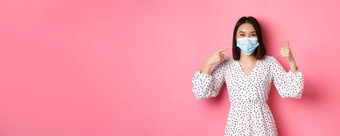 冠状病毒社会距离生活方式概念可爱的亚洲女人指出脸面具显示翘拇指支持预防措施科维德粉红色的背景