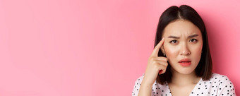 美皮肤护理概念头像可爱的亚洲女人盯着困惑指出手指头皱着眉头站粉红色的背景