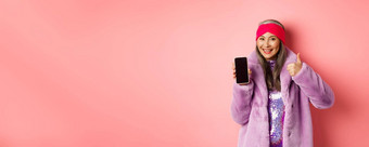 在线购物时尚概念时髦的亚洲高级女人显示空白移动屏幕竖起大拇指推荐互联网促销粉红色的背景