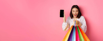 有吸引力的亚洲女人显示智能<strong>手机应用</strong>程序购物袋购买在线<strong>应用</strong>程序站粉红色的背景