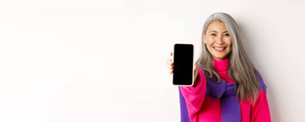 在线购物关闭现代亚洲<strong>高级</strong>女人扩展手移动电话显示空白智能手机屏幕微笑站白色背景