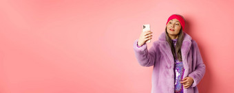 时尚概念时尚的亚洲高级女采取自拍智能手机摆姿势紫色的人造皮毛外套聚会，派对衣服站粉红色的背景