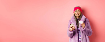 在线购物时尚概念微笑中年女人时尚的皮毛外套移动电话塑料信贷卡站快乐粉红色的背景