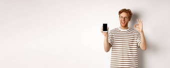 技术电子商务概念年轻的男人。红色的头发显示标志空白智能手机屏幕赞扬太棒了应用程序站白色背景