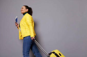 底视图女旅行者女人黄色的夹克蓝色的牛仔裤微笑走手提箱孤立的灰色的背景旅行持有空气票身份文档手