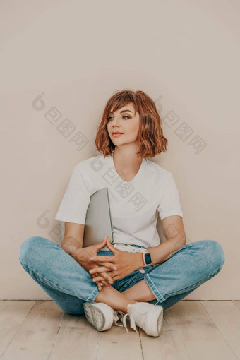 浅黑肤色的女人坐在地板上移动PC米色墙背景穿白色t恤牛仔裤白色运动鞋