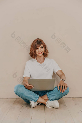 浅黑肤色的女人坐在地板上移动PC米色墙背景穿白色t恤牛仔裤白色运动鞋