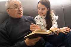 照片岁的爷爷漂亮的（外）孙女坐沙发拥抱保持房子检疫安全读有趣的书仙女演讲现代室内生活房间在室内