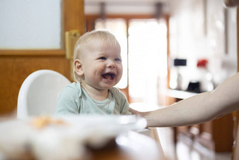 可爱的快乐的快乐婴儿婴儿男孩孩子微笑坐着高椅子餐厅表格厨房首页还是现在勺子美联储妈妈。