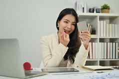 快乐的年轻的亚洲女人工人坐着工作桌子上应用使口红嘴唇