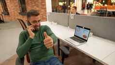 高加索人有胡子的男人。会说话的智能手机坐着桌子上现代共同办公空间商人相机显示拇指
