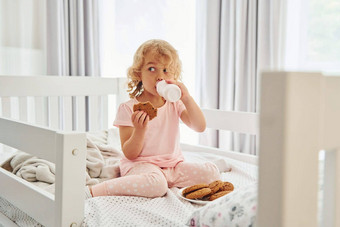 坐着床上吃<strong>饼干</strong>喝牛奶可爱的女孩<strong>休闲</strong>衣服在室内首页白天