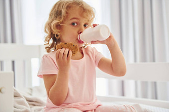 坐着床上吃饼干喝牛奶可爱的女孩休闲衣服在室内首页白天