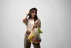 生态友好的积极的年轻的女人米色超大的t恤持有可重用的网棉花生态袋购物食品杂货白色背景
