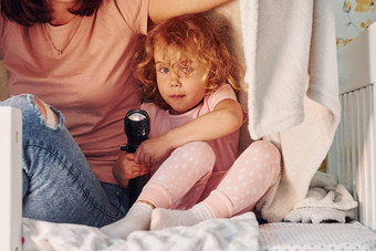 坐着毯子手电筒年轻的妈妈。女儿休闲衣服在室内首页