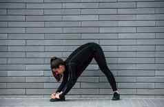 年轻的运动型女孩黑色的运动服装在户外伸展运动灰色的墙