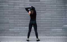 后视图年轻的运动型女孩黑色的运动服装站在户外灰色的墙