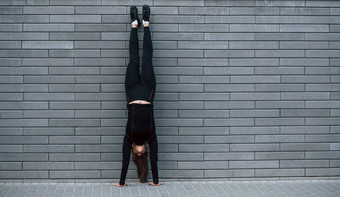 年轻的运动型女孩黑色的运动服装硬倒立练习在户外灰色的墙