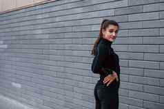 年轻的运动型女孩黑色的运动服装站在户外灰色的墙