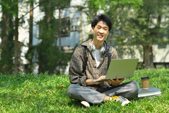微笑亚洲男人。学生移动PC绿色草前面大学建筑教育技术生活方式概念